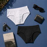 Two pairs of silkcut micro modal air men's briefs, one white and one black, arranged in a flat lay on a blue surface with sunglasses, a wallet, a key fob, and a book.