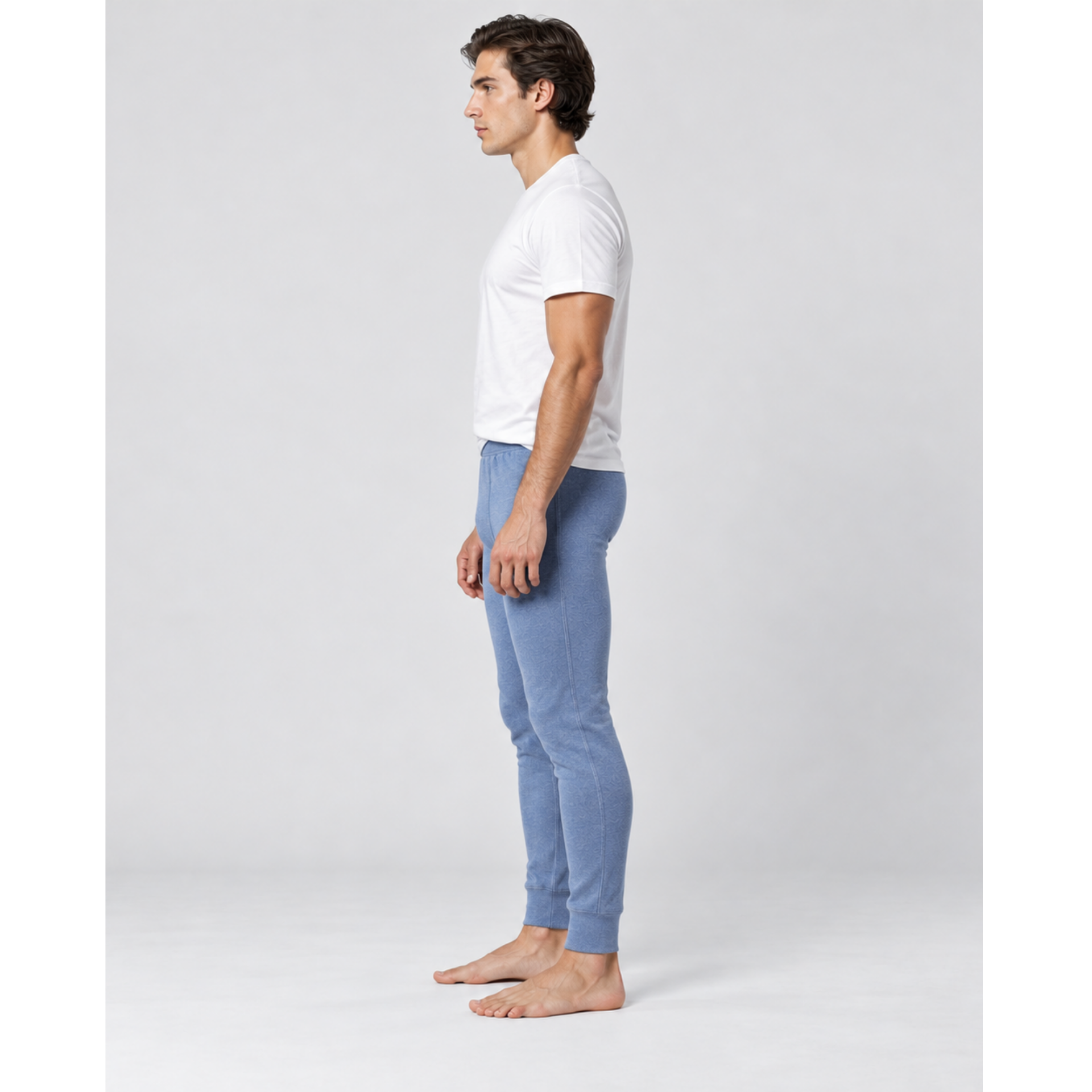 A man in profile wearing a white short-sleeve t-shirt and light blue silkcut thermal leggings for men standing barefoot against a light gray studio background.