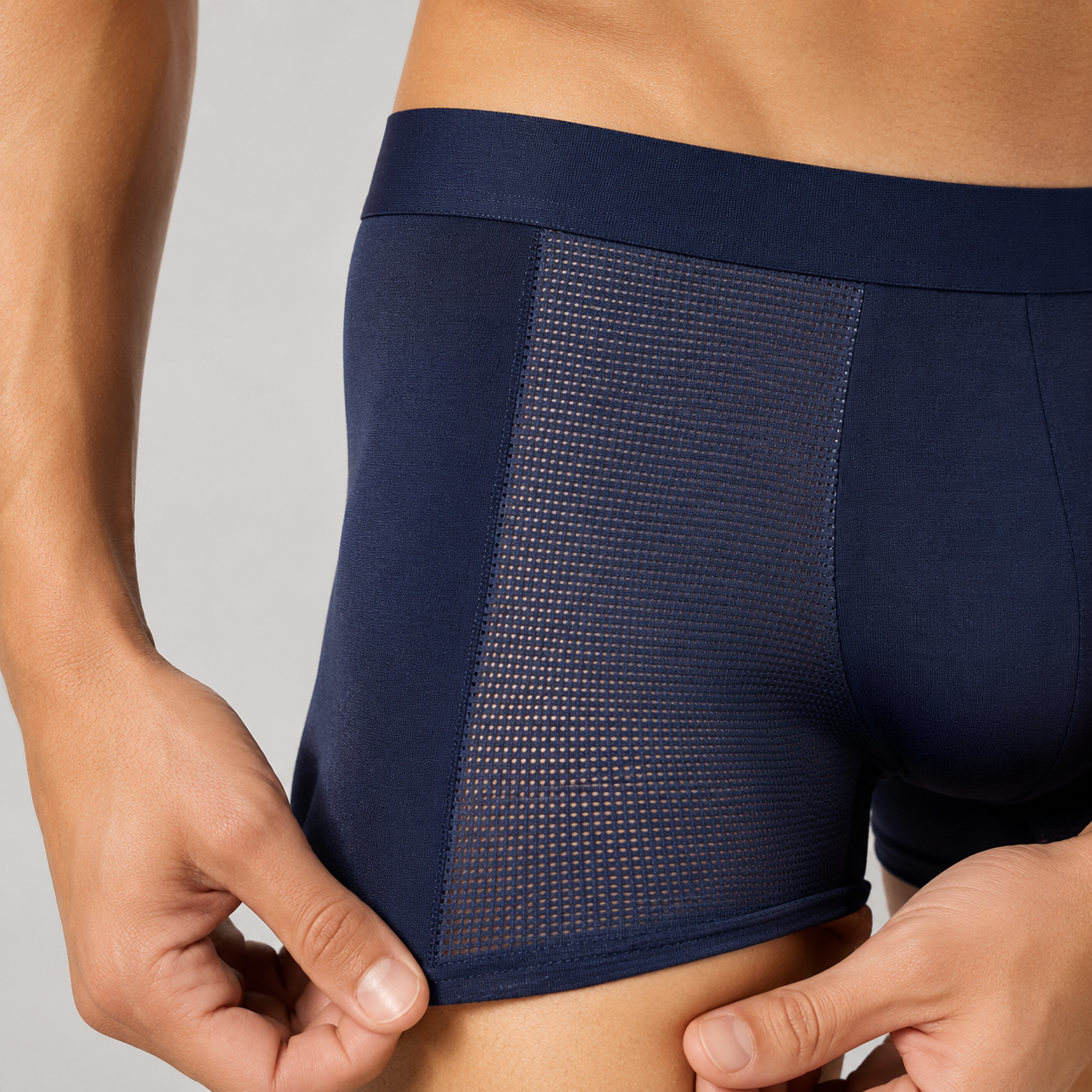 Close-up of a person wearing navy blue boxer briefs, showing the side and front mesh panel of the underwear being held slightly down by both hands against a light gray background.
